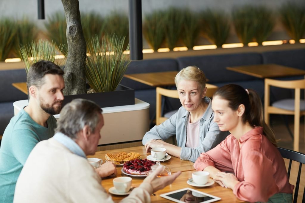 Four people talking over coffee and cake at a café table, a tablet in front of them, planning an emergency fund for self-employed people