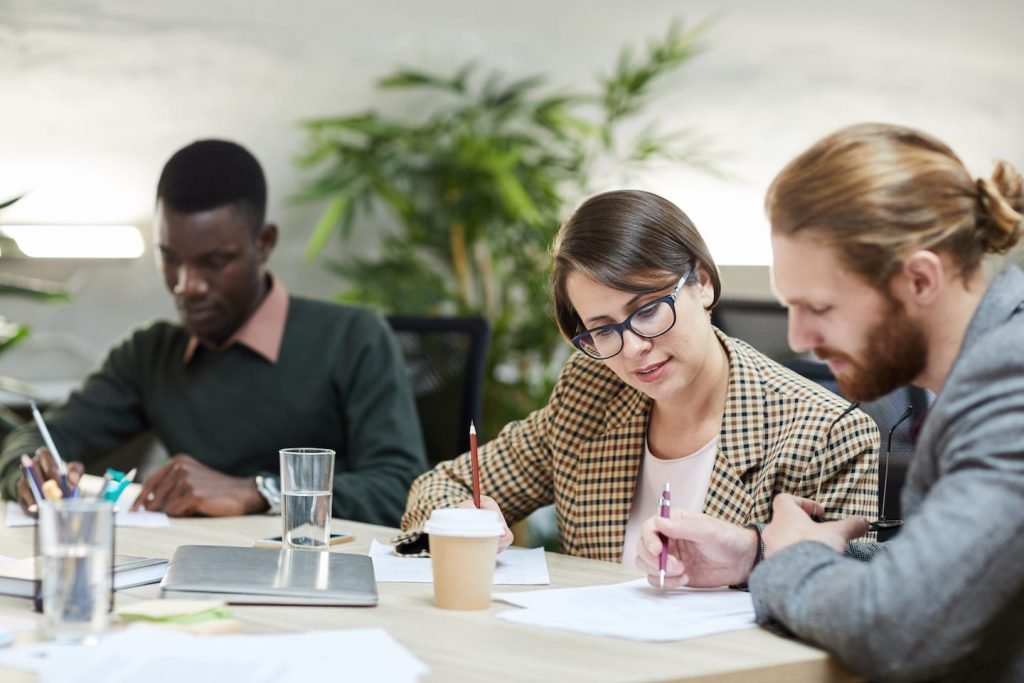 Three colleagues at a shared table, a woman in glasses going over papers with a coworker, planning to turn a side hustle into a business