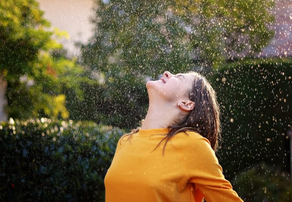 Woman in a yellow sweater tipping her face up to falling rain in a sunlit garden