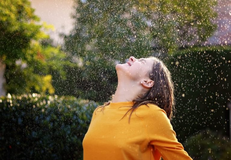 Woman in a yellow sweater tipping her face up to falling rain in a sunlit garden