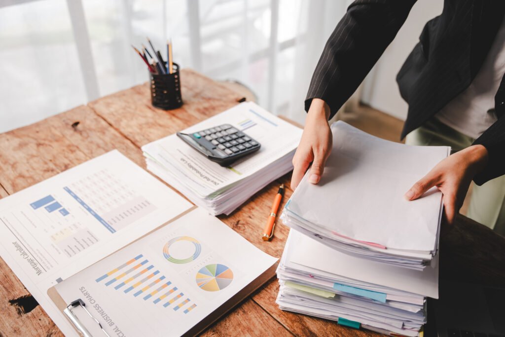 Hands sorting tall stacks of paperwork on a wooden desk with charts and a calculator