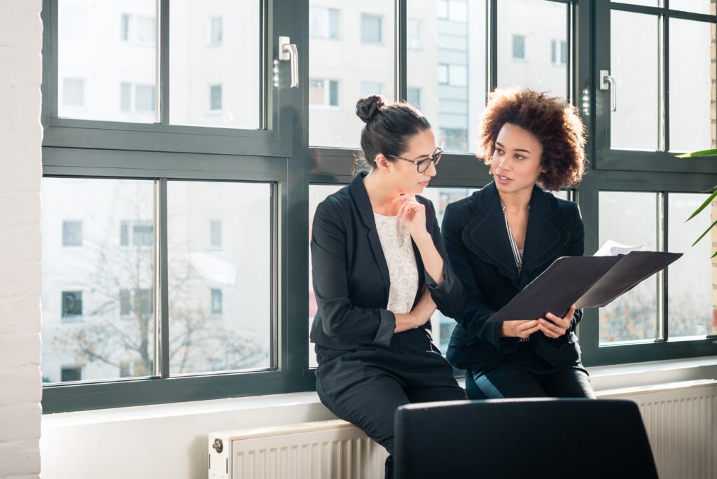 Two businesswomen talking by a window, practicing how to negotiate with clients for a higher rate