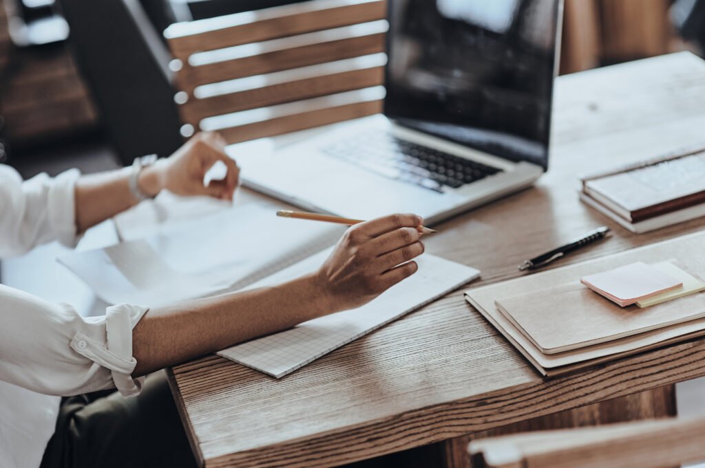 Woman's hands with a pencil and notepad at a wooden desk beside a laptop and notebooks
