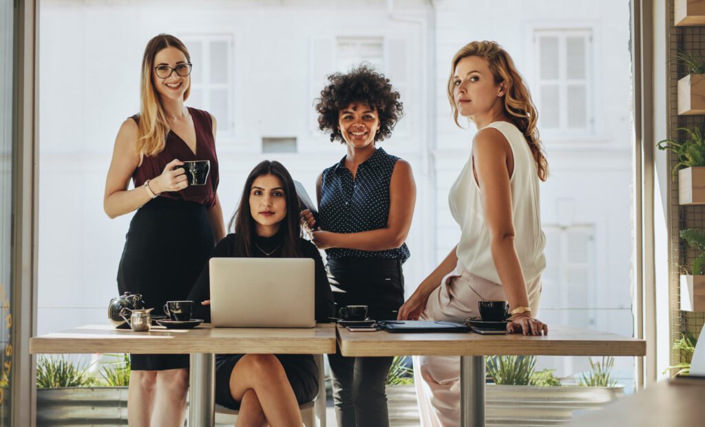 Four women posing around a table with a laptop and coffee cups, a team ready to cut business expenses without hurting growth