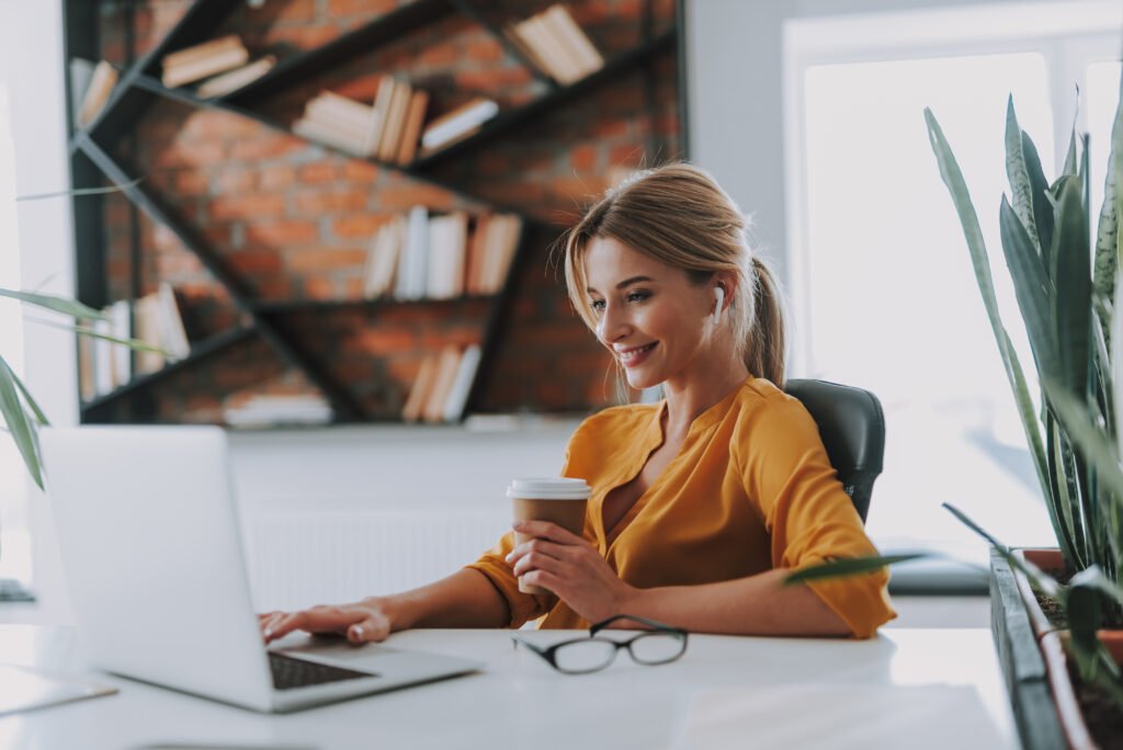 Smiling woman in a mustard blouse holding a coffee cup while working on a laptop