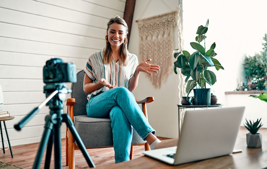 Smiling woman in an armchair talking to a camera on a tripod, plants and a laptop nearby, recording on a batch content creation day