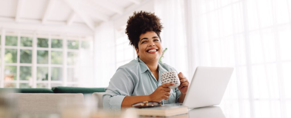 A smiling curly haired woman at her laptop, feeling good about how much she'll charge now.