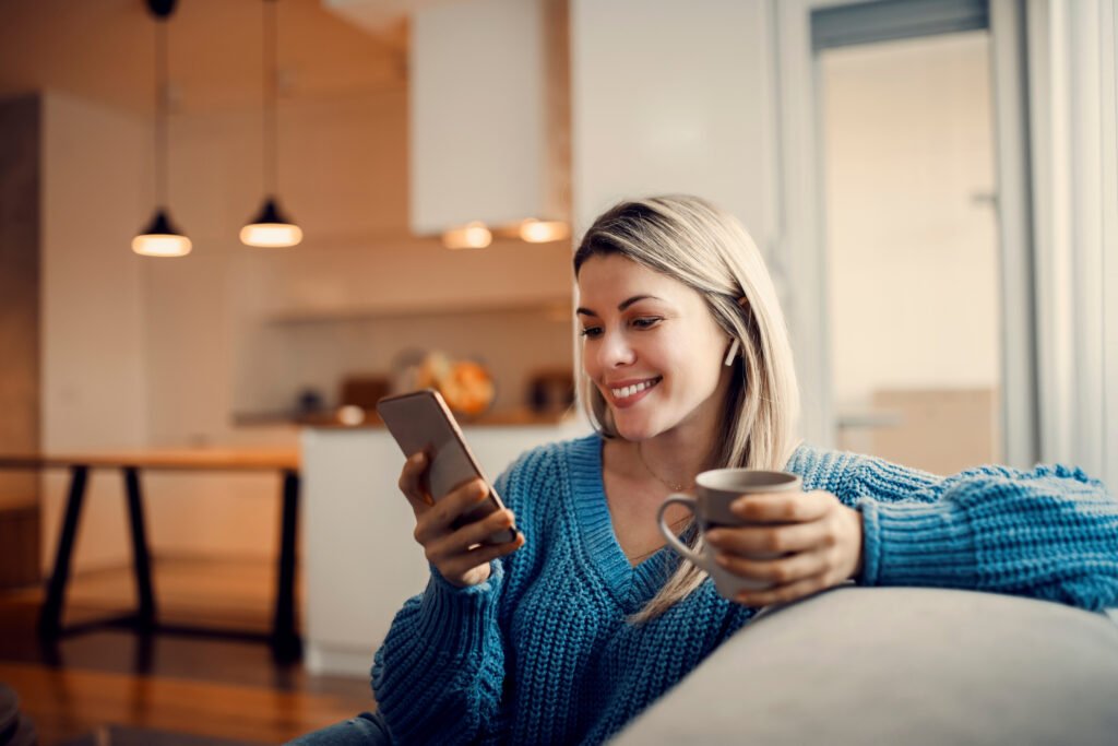 Smiling woman in a blue sweater on a sofa with her phone and a mug
