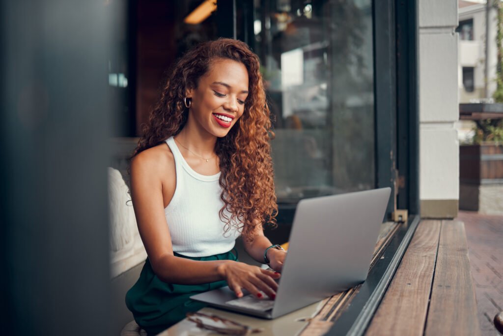 Smiling woman with long curly hair typing on a laptop at a café window counter