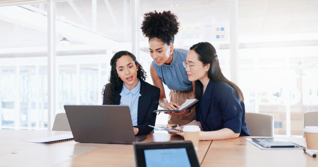 Three women discussing work around a laptop in a bright office, brainstorming ways to make more money in their business