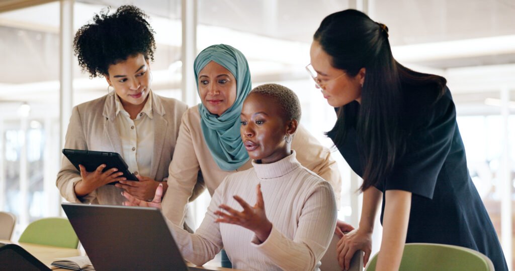 Four women gathered around a laptop in an office, talking through how to build multiple revenue streams