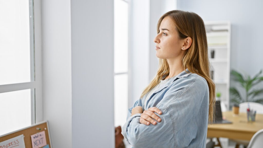 Woman with arms crossed looking out an office window, deep in thought