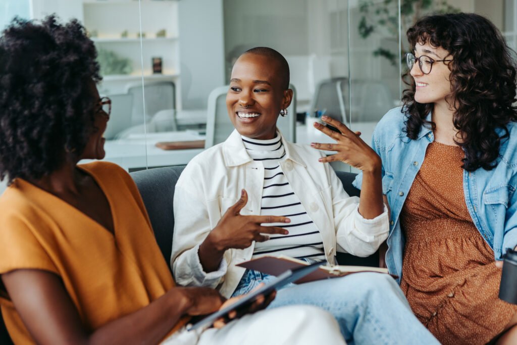 Three women talking and smiling on a sofa in a bright office