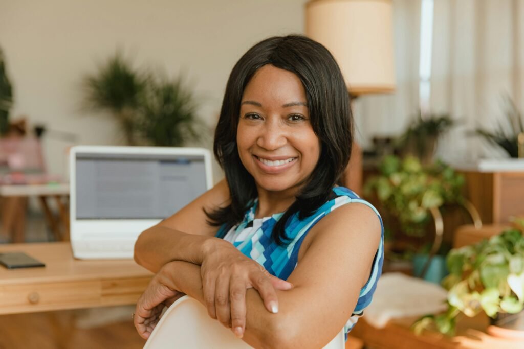 Smiling woman with a dark bob leaning on a chair, laptop and plants behind her