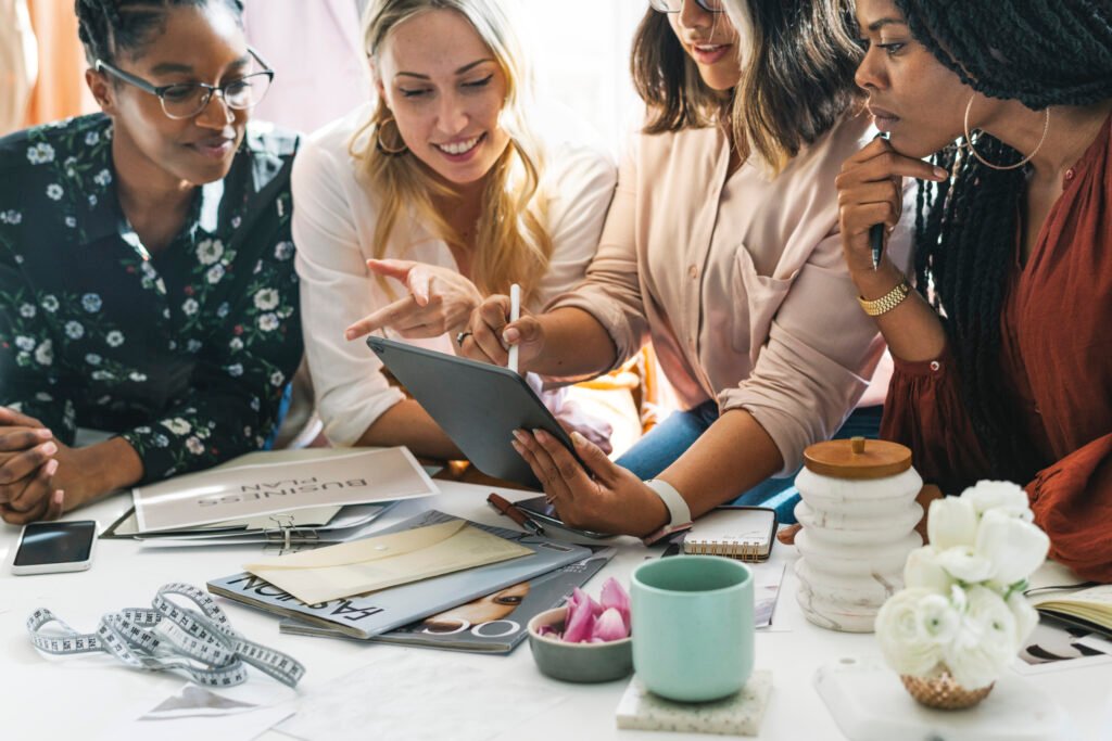 Four women gathered around a tablet at a table with magazines, a tape measure and a mug, learning to manage business finances