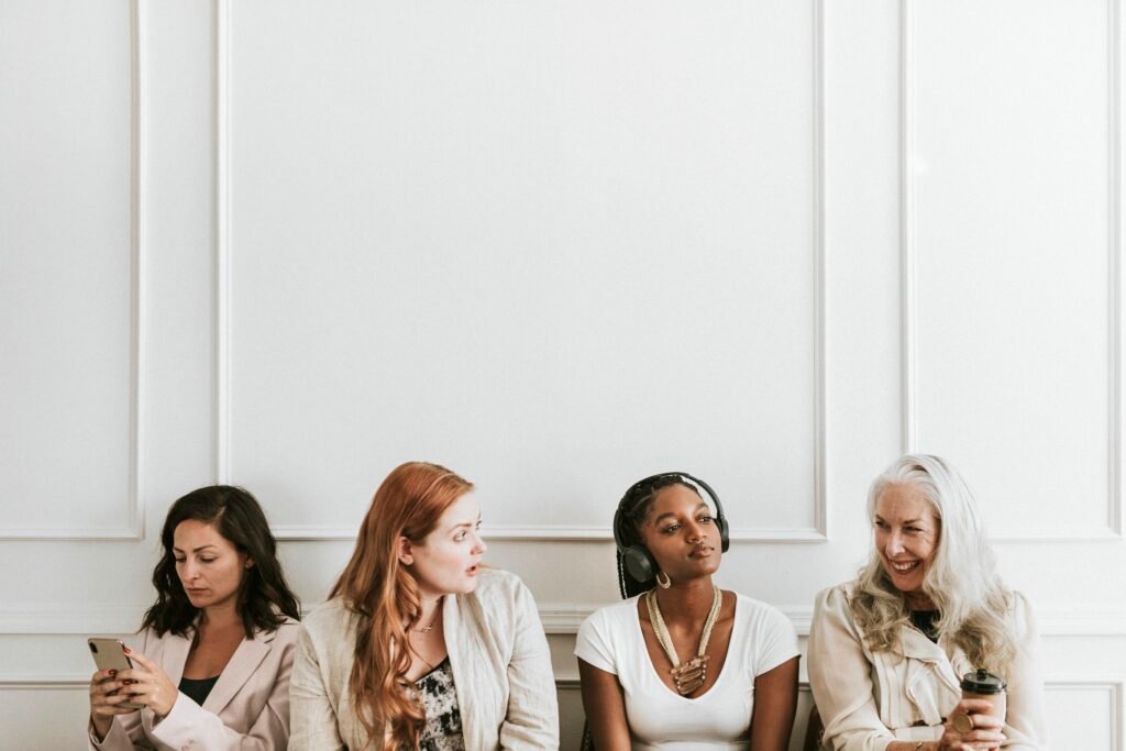 Four women of different ages sitting in a row against a white wall, one on her phone, each facing her own money traps