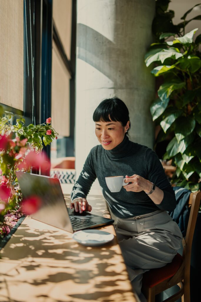 A woman working at a laptop at a sunny desk full of plants
