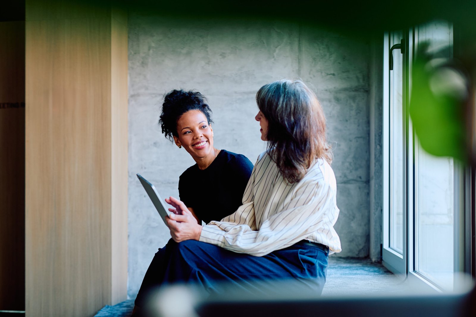 Two women sitting by a window in a warm conversation, one holding a tablet