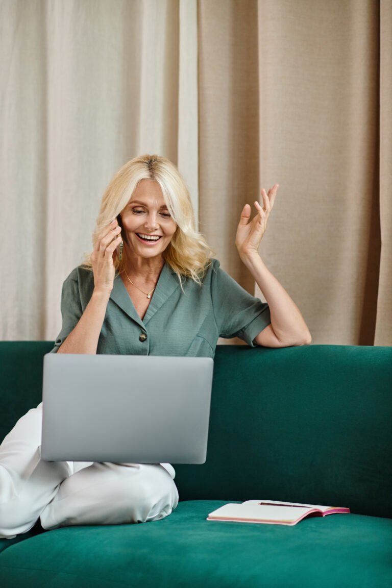 Laughing blonde woman on a phone call with a laptop on a green velvet sofa