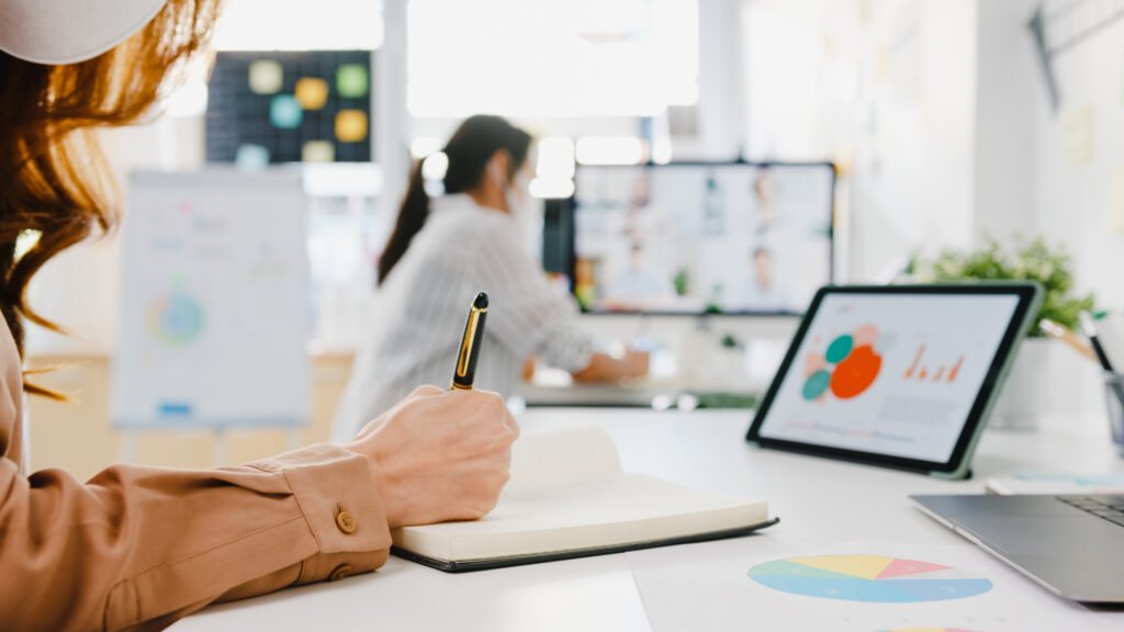 Woman taking notes beside a tablet showing charts, a colleague on a video call behind her, learning to write content for AI search