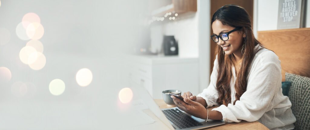Smiling woman in glasses looking at her phone with her laptop open at home