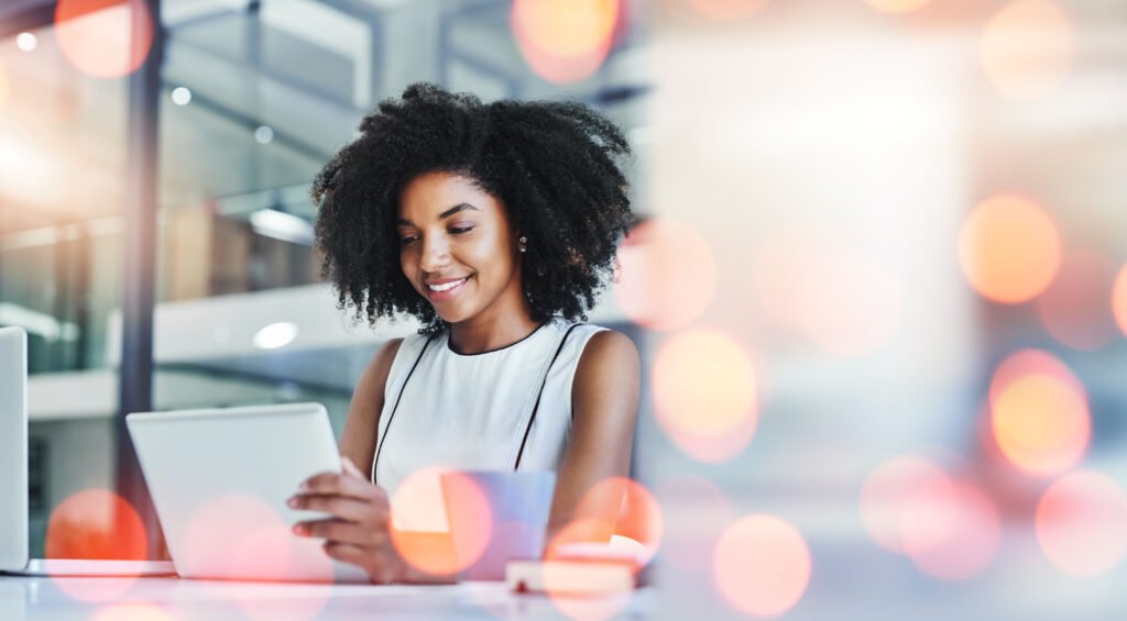 A young woman with curly hair smiling at a tablet at her desk, bright bokeh lights behind her, working out how to get her business on ChatGPT.