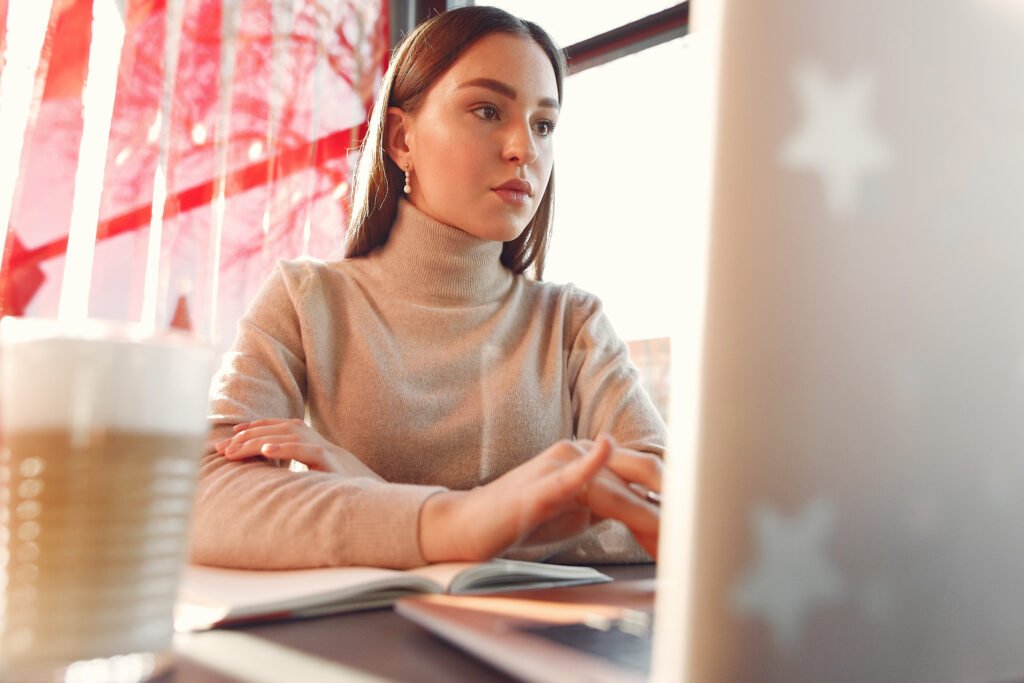 Woman in a beige turtleneck typing on a laptop beside an open notebook and a latte