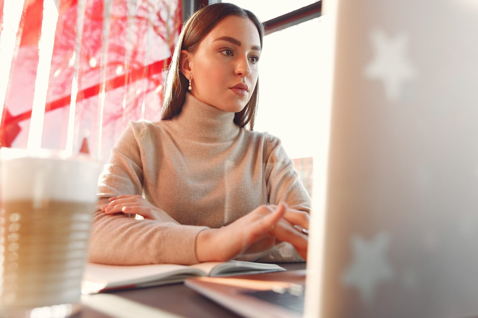 Woman in a beige turtleneck typing on a laptop beside an open notebook and a latte