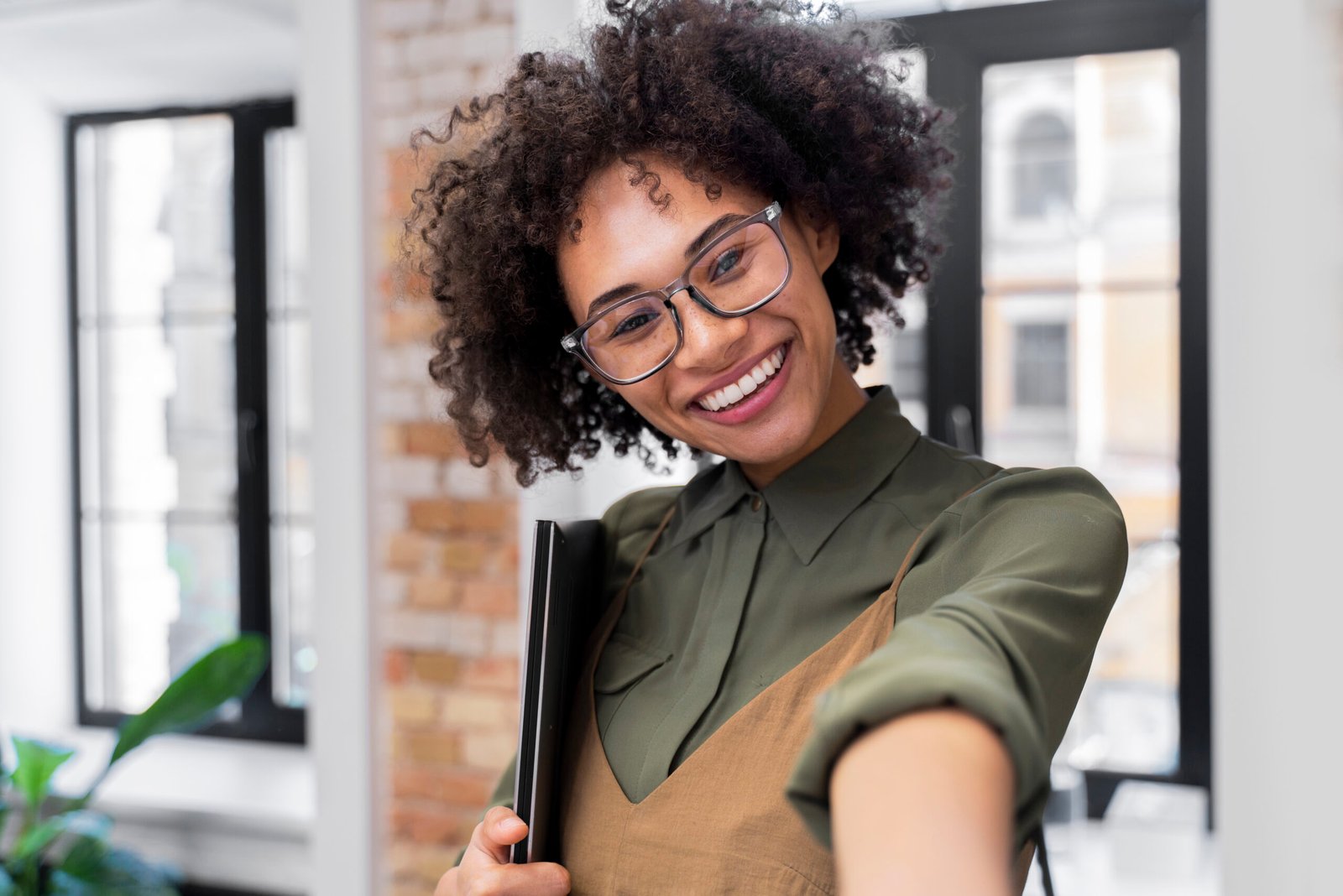 Smiling woman with curly hair and glasses holding a black folder, leaning toward the camera