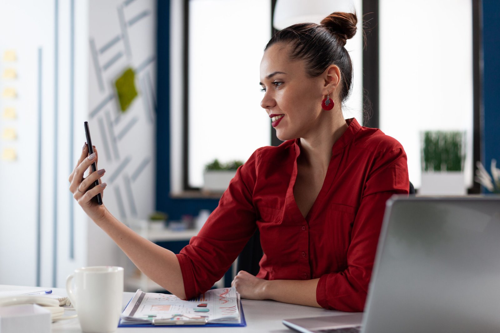 Woman in a red blouse smiling into her phone held at arm's length at her desk