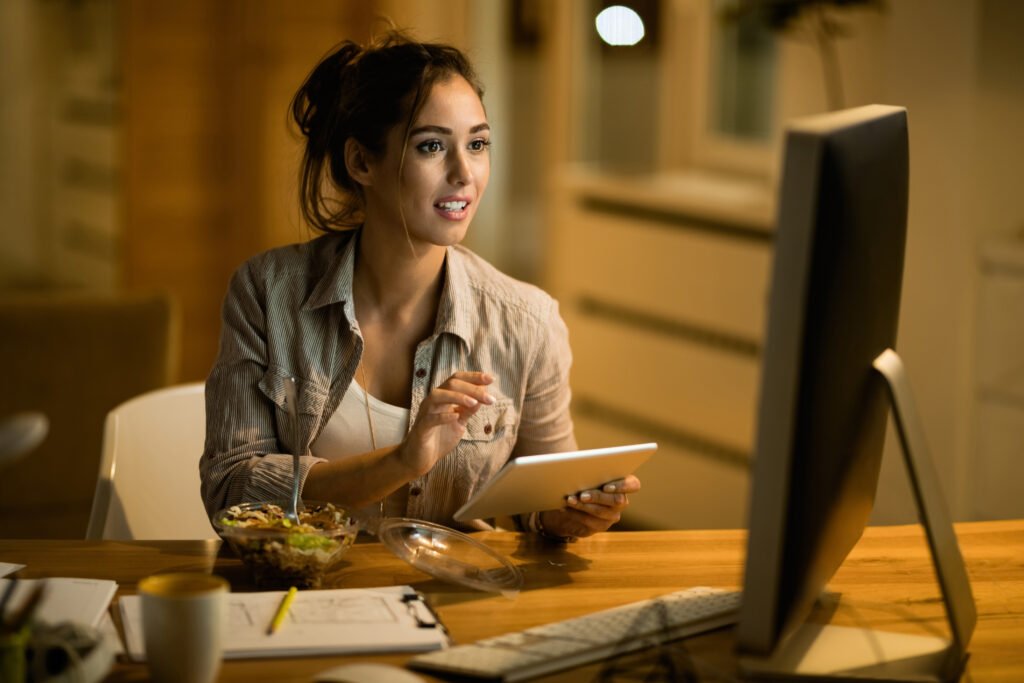 A young woman holding a tablet while checking her desktop monitor at night, mid-build and working out how long to make an app with AI.