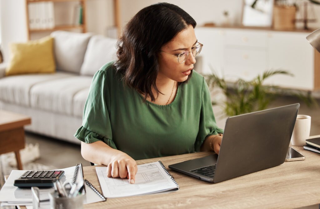 Woman in glasses and a green blouse studying a notebook beside her laptop and calculator, mapping out her influencer income streams