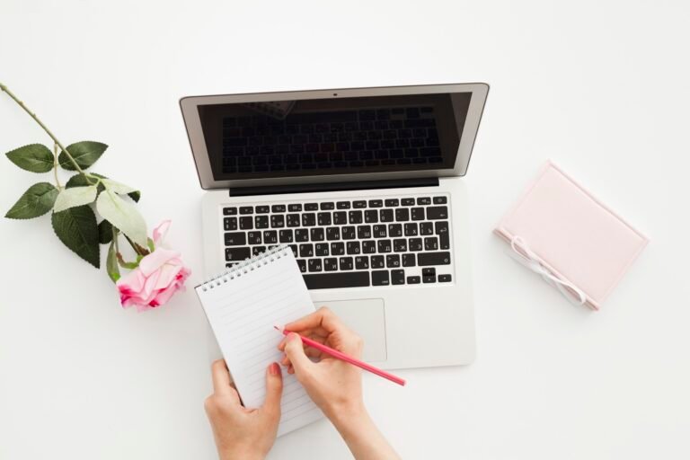 Overhead view of hands writing on a notepad beside a laptop and pink roses on a white desk