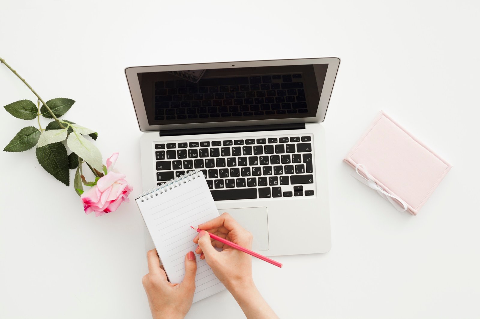 Overhead view of hands writing on a notepad beside a laptop and pink roses on a white desk