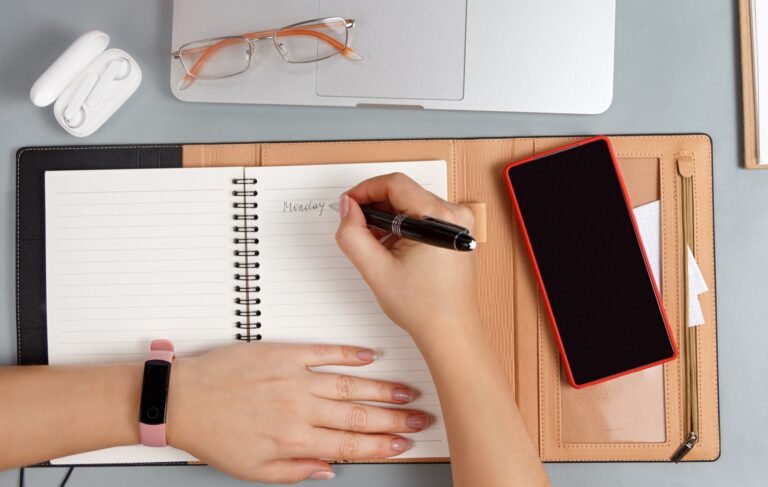 Overhead view of hands writing Monday in a planner beside a laptop, glasses and a red phone
