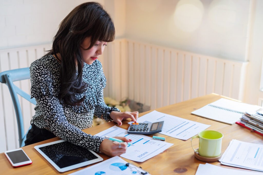 Young woman with a calculator highlighting printed charts at a pale wooden table, a tablet beside her, checking the math by hand before she builds a calculator for her website