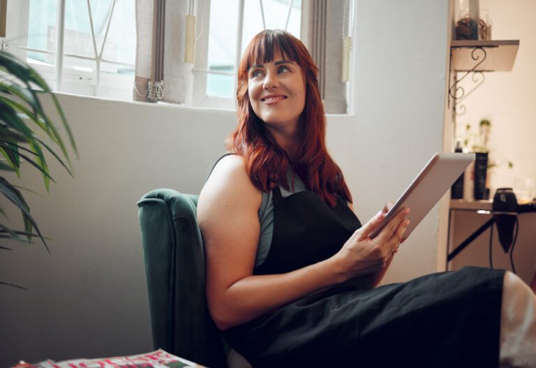 Smiling red-haired woman in a green velvet armchair with a tablet, gazing toward the window