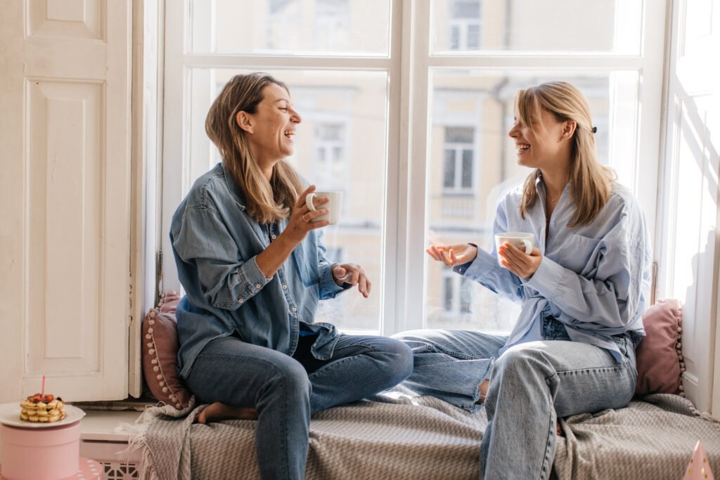 Two women laughing over coffee on a sunny windowsill