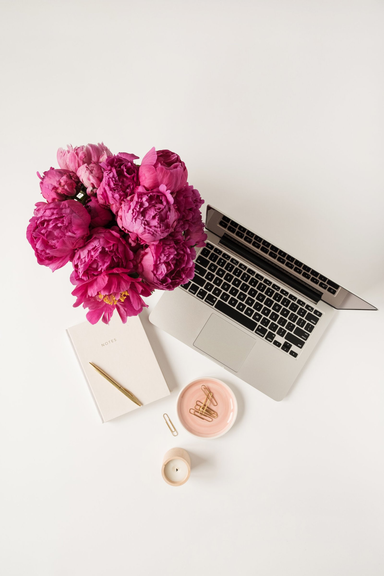 A laptop, notebook and pink peonies on a white desk
