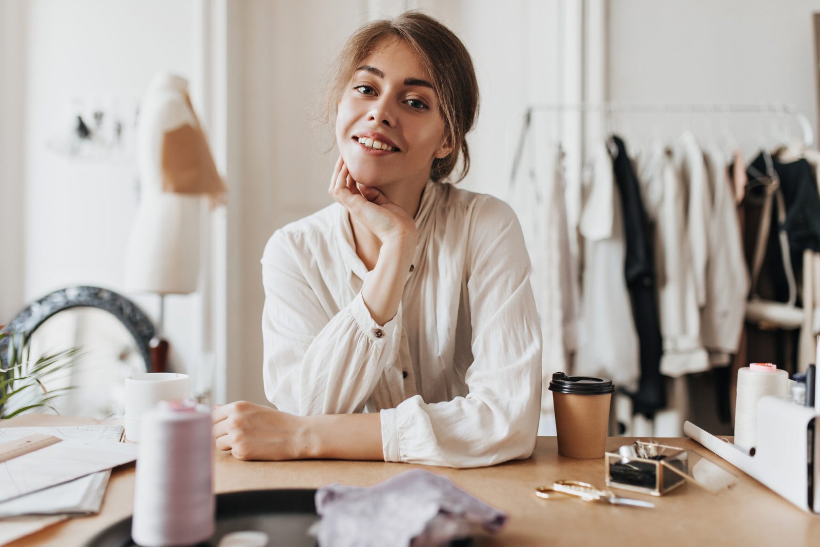 Smiling woman in a cream blouse at a sewing table, dress form and clothing rail behind