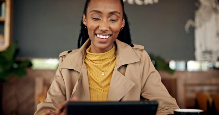 Smiling woman with braids in a trench coat looking down at her tablet in a café