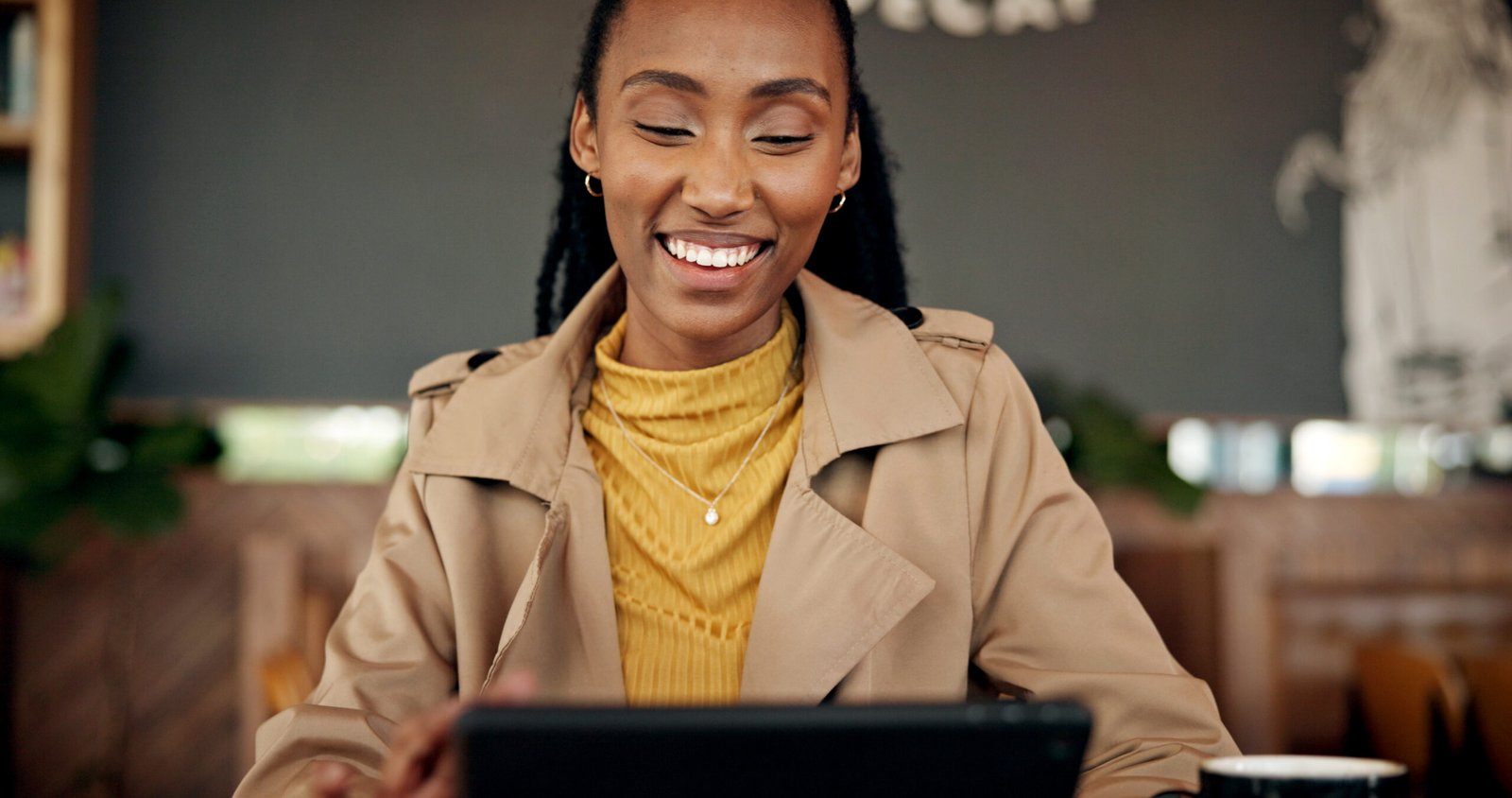 Smiling woman with braids in a trench coat looking down at her tablet in a café