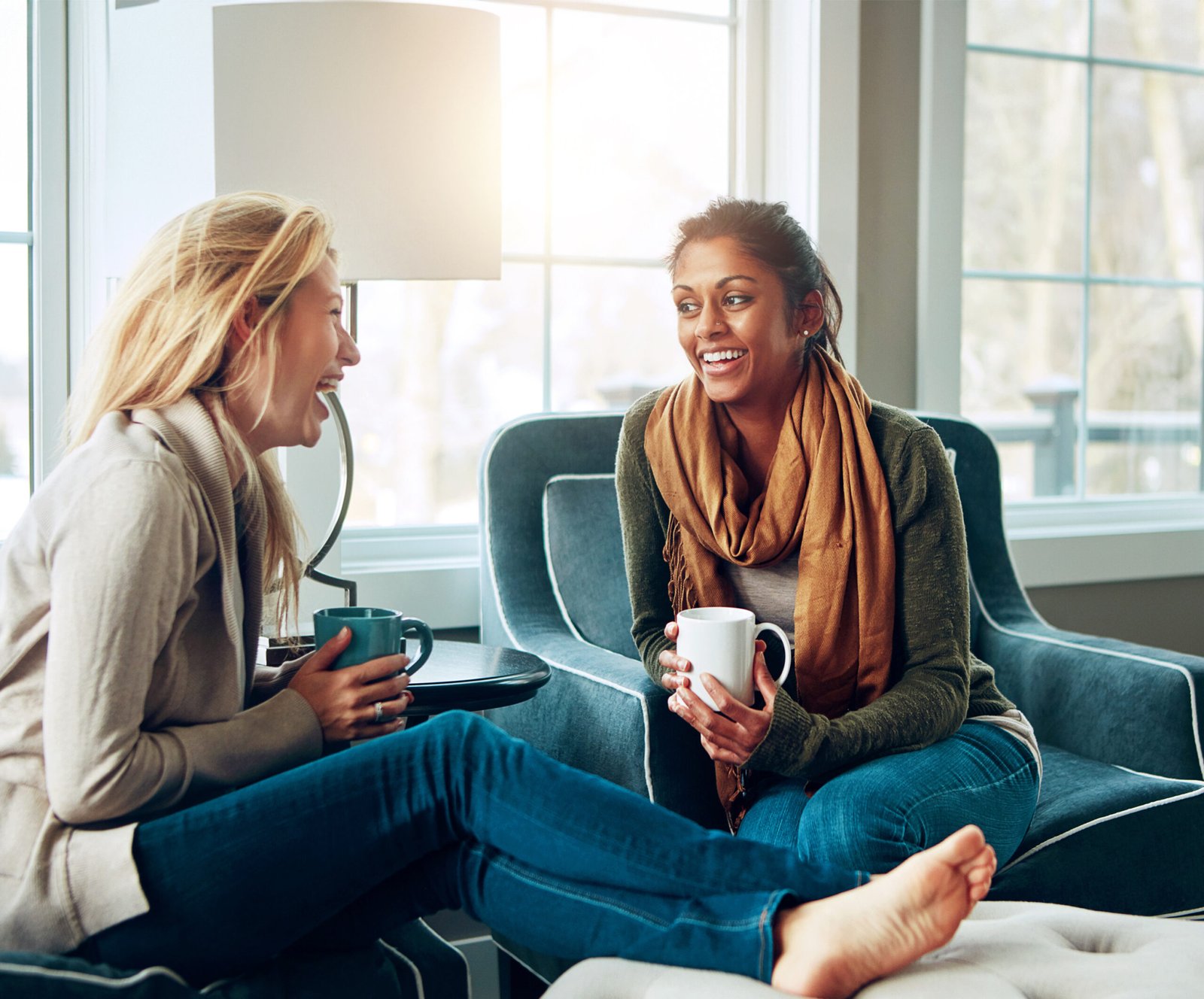Two women laughing over mugs of coffee in armchairs by a bright window