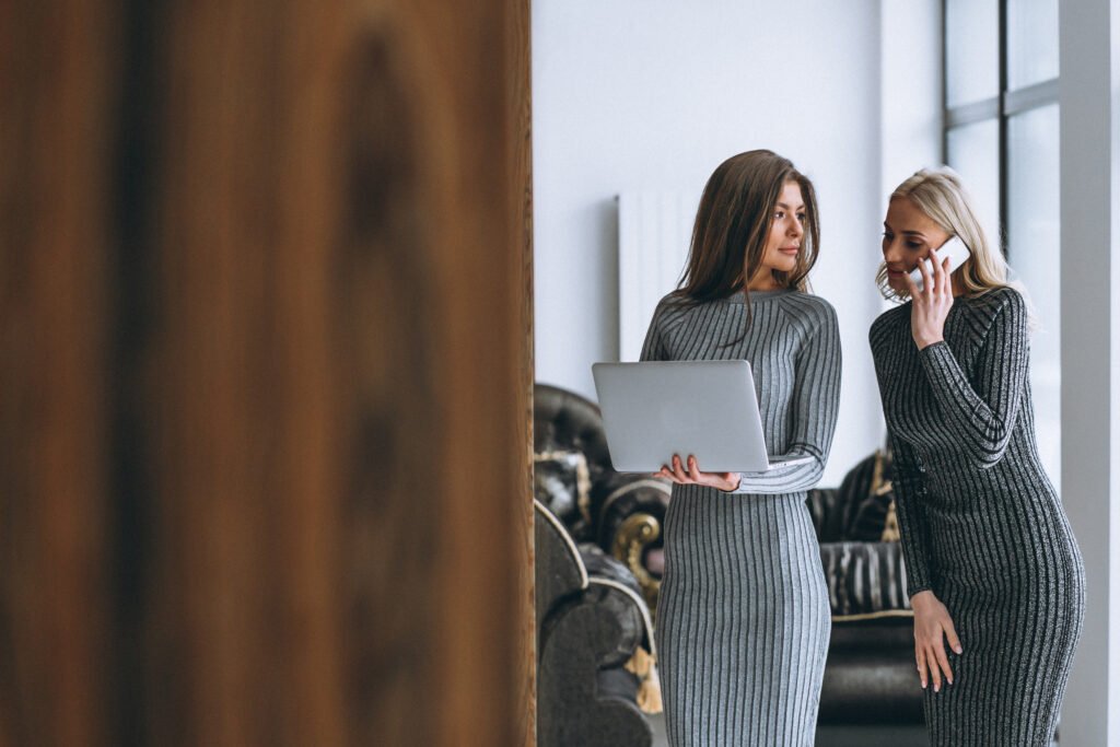 Two women in grey knit dresses by a window, one holding a laptop and one on the phone, talking through the 50/30/20 rule