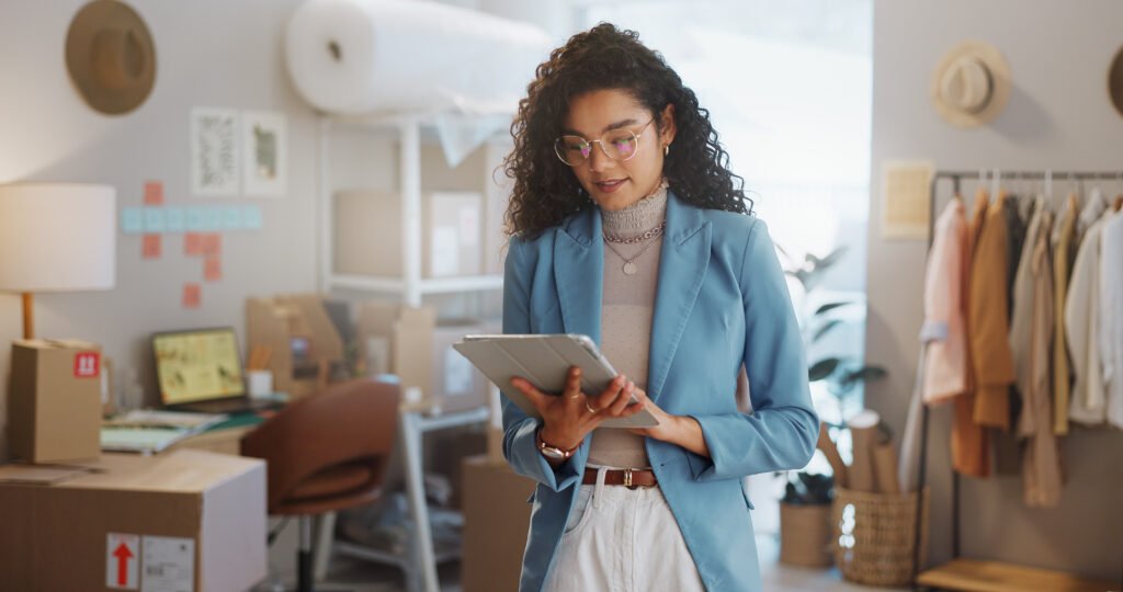Woman in a blue blazer checking a tablet in a boutique stockroom full of boxes