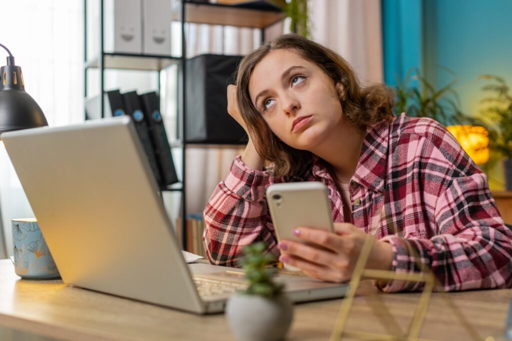 Young woman rolling her eyes at her phone while working at a laptop in a home office