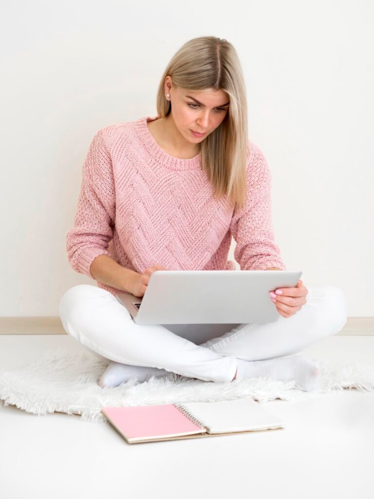 A blonde woman sitting cross legged on the floor with her laptop, happy running a business she doesn't have to scale.