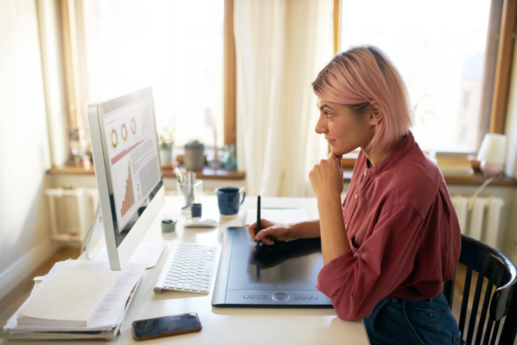 Woman with a pink bob using a stylus and drawing tablet at a computer showing charts