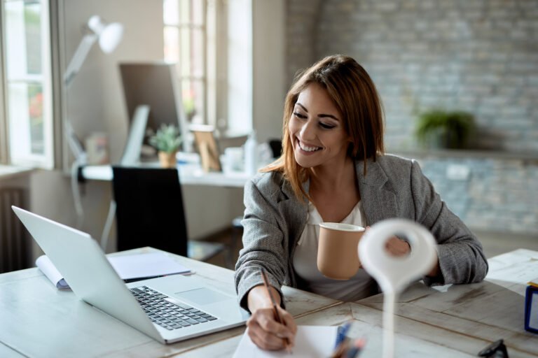 Smiling woman with a mug writing notes beside her laptop at a bright office desk